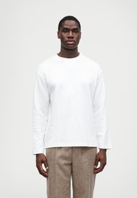 Mladić stoji licem prema naprijed, noseći bijelu košulju dugih rukava i bež teksturirane hlače, protiv jednostavne svijetle pozadine.