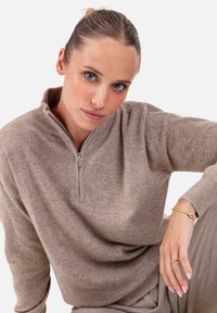 Pull marron clair à fermeture éclair avec col côtelé et manches longues, offrant une texture douce et une coupe décontractée, associé à un pantalon beige lisse.