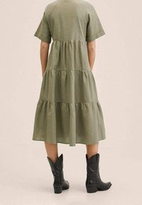 Olivgrünes, gestuftes Kleid aus weichem Stoff, mit kurzen Ärmeln, lässigem Schnitt und dezenten Raffung in der Taille, kombiniert mit schwarzen Stiefeletten.