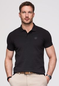 Mann mit kurzen braunen Haaren, der ein schwarzes Poloshirt, beige Hose, braunen Gürtel, Uhr und Armbänder trägt, steht mit den Händen in den Taschen.