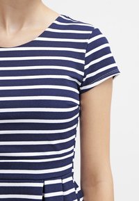 Tummansininen ja valkoinen raidallinen mekko, jossa on lyhyet hihat, pyöreä pääntie ja pleatattu vyötärösuunnittelu. Pehmeä, tekstuurinen kangas.