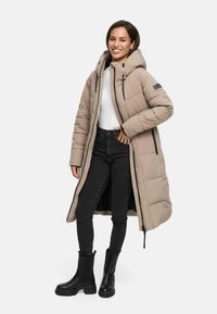 Lange beige pufferjas met een capuchon, rits aan de voorkant en zwarte accenten. Draag het over een witte top, in combinatie met zwarte jeans en zwarte laarzen.