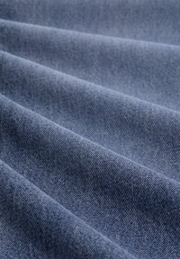 Denimová tkanina ve středně modré barvě, s jemnou strukturou a viditelným herringbone vzorem, složena tak, aby ukázala hloubku a strukturu.