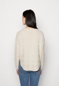 Pull en maille beige avec texture côtelée, col rond et manches raglan. Présente un ourlet asymétrique et des fentes latérales. Porté avec un jean bleu.