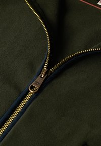 Oliwkowa kurtka o gładkiej fakturze, z złotym zamkiem błyskawicznym i granatowymi wykończeniami. Zamek błyskawiczny ma zawieszkę z logo "SUPER DRY".