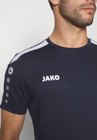 Maglietta sportiva blu navy con accenti grigi, con il logo "JAKO" in bianco. Texture liscia, maniche corte con dettaglio circolare sulle spalle.