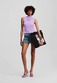 Lila brezrokavni top z logotipom, kavbojke s frayed robom, črna torba za čez ramo z grafičnim detajlom in svetlo vijolične salonarje na odprto nogo.