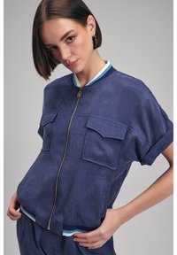 Mörkblå bomberjacka med korta ärmar, dragkedjestängning, två bröstfickor och randig ribbad krage. Lättviktigt tyg med en texturerad yta.