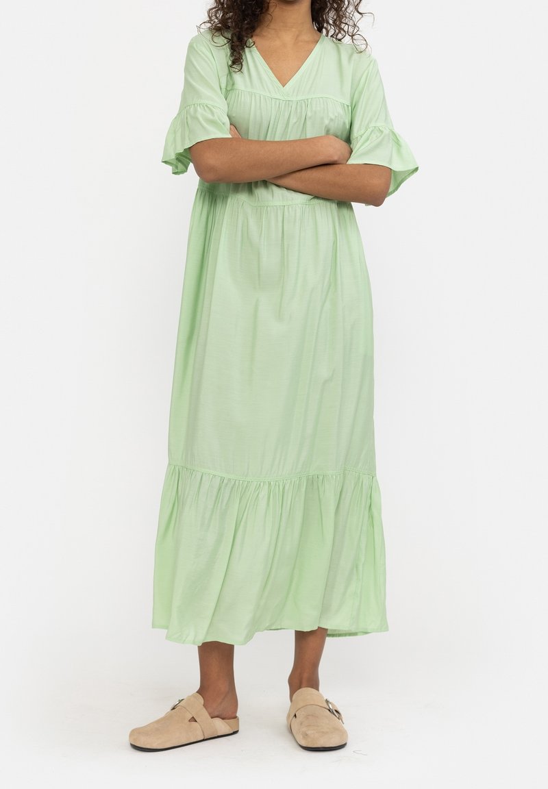 Robe longue fluide vert clair avec un col en V et des manches à volants superposés, présentant une coupe ample et un tissu doux et texturé. Portée avec des sabots beiges.