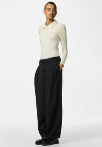 Kremowe, prążkowane polo z długim rękawem i trzema guzikami, zestawione z luźnymi, czarnymi plisowanymi spodniami oraz czarnymi mokasynami.