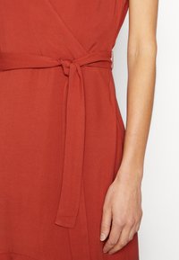 Vestido em tom ferrugento com design de envoltório, apresentando um nó na cintura. O tecido parece suave e leve, destacando linhas limpas e simplicidade.