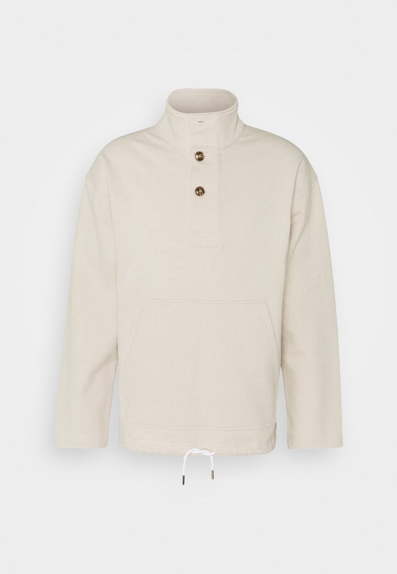 Beżowy sweter wykonany z miękkiego materiału. Posiada wysoki kołnierz, zapinany na trzy guziki, oraz przednią kieszeń z regulowanym troczkiem u dołu.