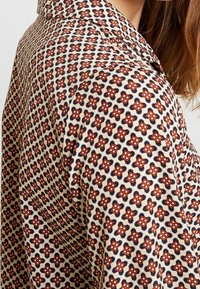 Camicia a maniche lunghe con sfondo crema e un motivo geometrico ripetuto in rosso e blu navy, dotata di colletto classico e texture in tessuto morbido.