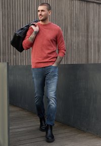 Homme tatoué portant un pull de couleur rouille et un jean, marchant en extérieur avec un sac noir sur l'épaule.