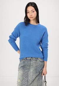 Modra pletena jopica z dolgimi rokavi in okroglo ovratnikom; nosi se z teksturiranimi, svetlo sivo sijočimi jeans hlačami.