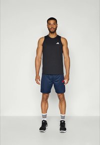 Atletische man die rechtop staat in een zwart mouwloos Adidas shirt, marineblauwe shorts, gestreepte witte sokken en zwarte Adidas sneakers op een effen achtergrond.
