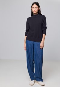 Dunkelgrauer Strickpullover mit hohem Kragen, kombiniert mit lockeren blauen Denimhosen und weißen Sneakers. Schlichtes und strukturiertes Design.