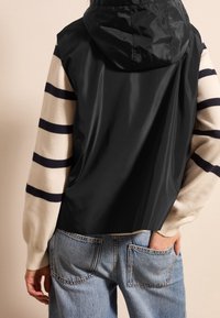 Black hooded vest over a cream and navy striped sweater; features a smooth, shiny finish and relaxed fit; denim jeans visible below.