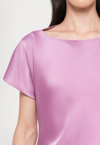 Femme portant un chemisier lavande en soie brillant à manches courtes, montrant le décolleté et le haut du bras sur un fond clair.