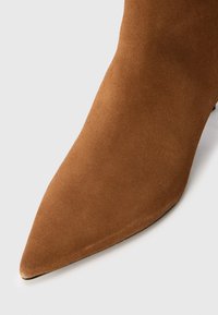 Stivale alla caviglia in suede marrone con punta affilata e texture liscia, caratterizzato da un design elegante e dettagli minimi.