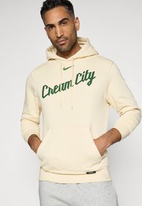 Hoodie v kremni barvi, izdelan iz mehkega blaga, z zelenim grafičnim napisom "Cream City" spredaj, nastavljivimi vrvicami in sprednjim žepom.