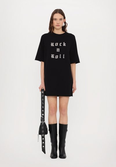 Vestido camisero negro de gran tamaño con un gráfico de "Rock n Roll" en plateado. Combinado con botas de cuero negras hasta la rodilla y un cinturón negro con tachuelas.