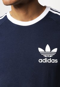 Tmavě modré bavlněné tričko s bílým kulatým výstřihem, třemi bílými pruhy na ramenou a bílým logem Adidas trefoil na hrudi.