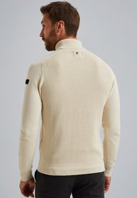 Hellbeige Strickpullover mit einem strukturierten Muster, hohem Kragen und gerippten Bündchen. Verfügt über ein kleines Logo am hinteren Halsausschnitt und schwarze Akzente.
