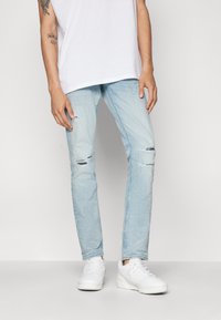 Vaqueros de denim azul claro con corte slim, que presentan detalles desgastados en las rodillas. Combinados con zapatillas blancas y una camiseta blanca básica.
