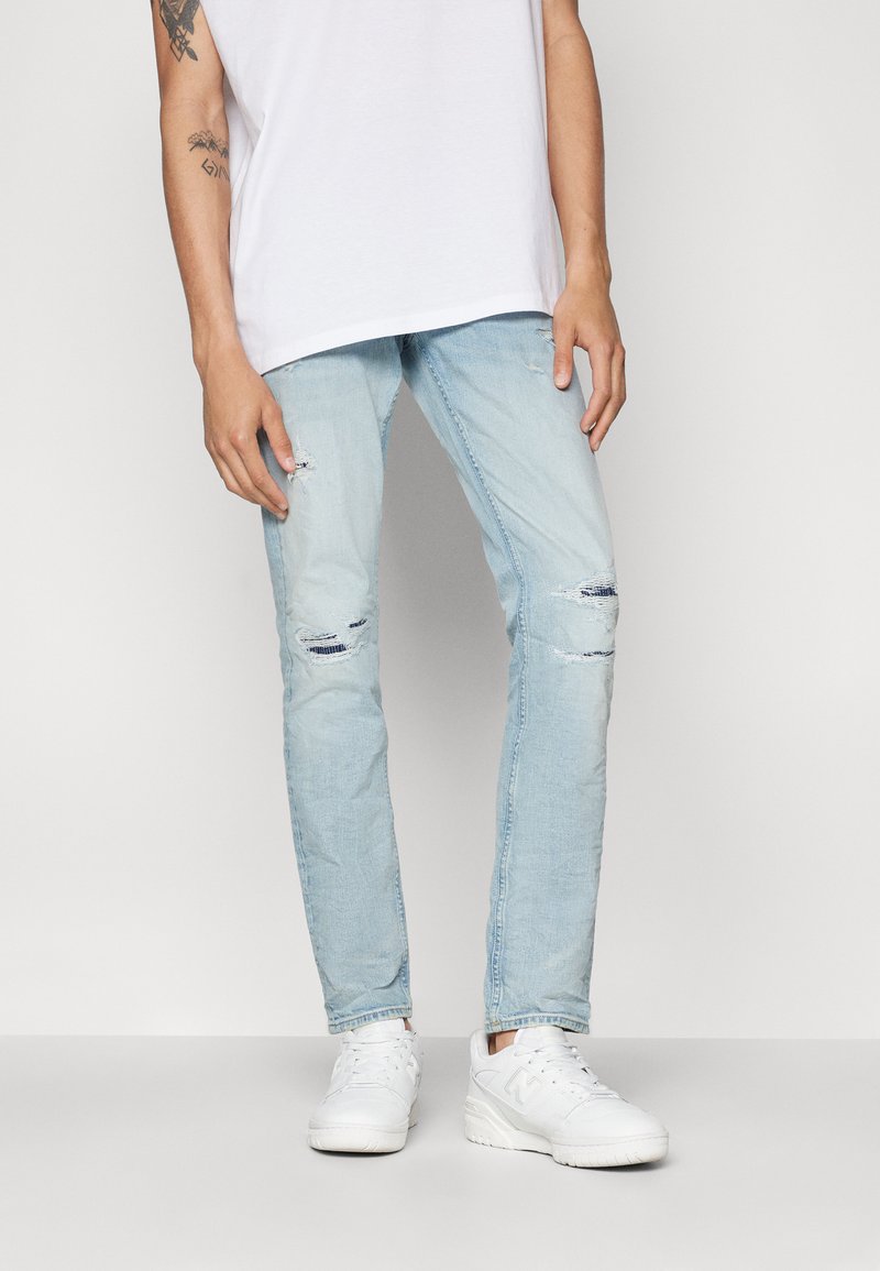Vaqueros de denim azul claro con corte slim, que presentan detalles desgastados en las rodillas. Combinados con zapatillas blancas y una camiseta blanca básica.