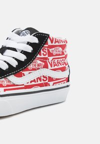 Baskets montantes avec un dessus en tissu rouge arborant un motif blanc "VANS", des accents en suède noir et des lacets blancs. Semelle en caoutchouc avec une bande noire.