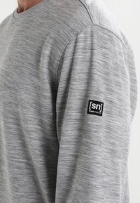 Camisa gris de manga larga con textura jaspeada, que presenta un pequeño parche del logo negro [sn] en la manga. Diseño de cuello redondo sencillo.