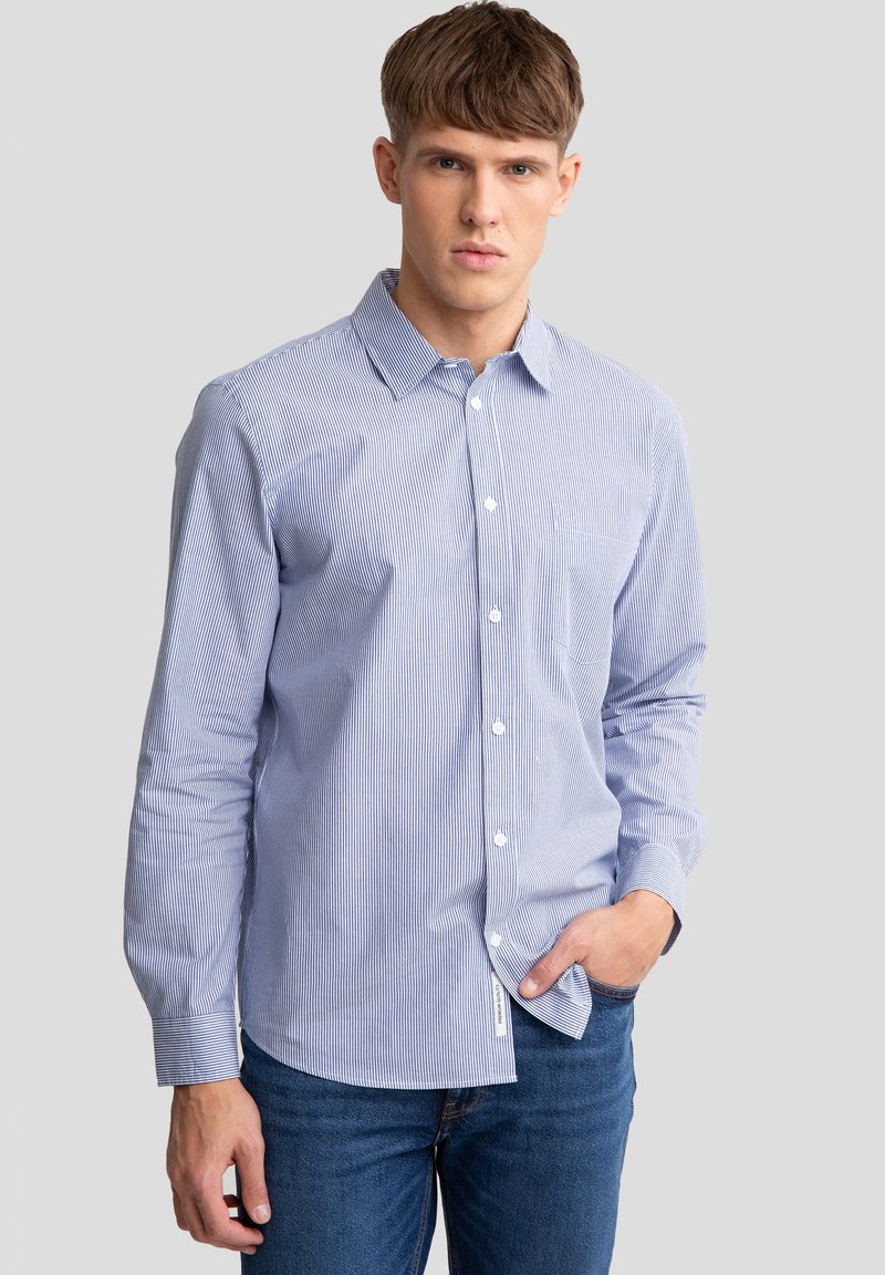Camicia a righe blu e bianche, con bottoni, maniche lunghe e colletto; presenta una tasca sul petto e orlo diritto. Indossata con jeans blu.