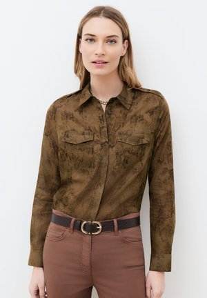 Mujer con cabello largo y rubio que lleva una camisa marrón con estampado de serpiente abotonada, pantalones marrones y un cinturón oscuro con hebilla dorada.