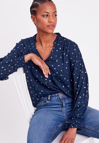 Marinebluse mit V-Ausschnitt und gerafftem Ausschnitt. Verfügt über ein dezentes Muster aus kleinen weißen Rhomben. Lange Ärmel mit Raffung an den Bündchen.