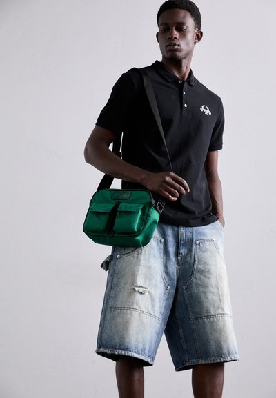 Grønn nylon crossover-veske med justerbar stropp, som har to fremre lommer og et svart logo-patch. Båret over en svart polo-skjorte og denimshorts.