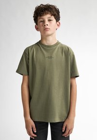 Camiseta de algodón verde oliva con mangas cortas, cuello redondo y un pequeño logo negro en el pecho. Combinada con jeans oscuros.