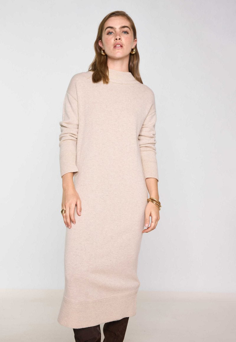 Abito in maglia beige con collo alto, maniche lunghe e polsini a costine. Presenta una silhouette dritta e un orlo che arriva fino a metà polpaccio.