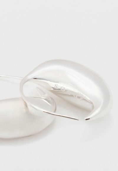 Primer plano de un pendiente de aro plateado torcido con superficie lisa y nombre de la marca grabado en el interior de la curva sobre un fondo blanco.