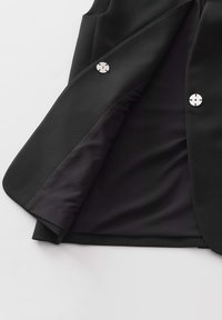 Schwarzer Blazer mit glatter Außenseite, silbernen Knopfdetails und einem kontrastierenden dunklen Futter, das ein sauberes, modernes Design präsentiert.