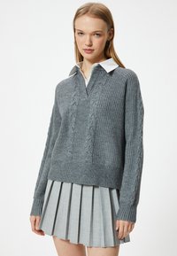 Grå stickad tröja med V-hals och flätmönster, kombinerad med en plisserad ljusgrå kjol. Har långa ärmar och ribbade muddar.