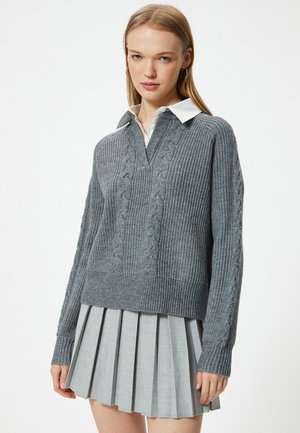 Suéter de punto gris con cuello en V y patrón de trenzas, combinado con una falda plisada gris claro. Presenta mangas largas y puños acanalados.