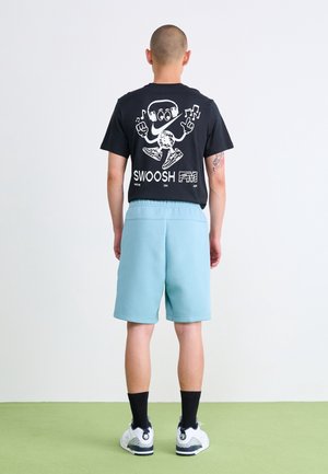Sort bomulds T-shirt med hvidt tegneserieagtigt grafisk motiv og tekst på ryggen, parret med lyseblå shorts og hvide sneakers. Grøn gulvbaggrund.