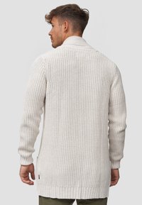 Cardigan lungo di colore beige chiaro, realizzato in maglia con trama tessuta. Presenta polsini e orlo a costine, colletto a scialle e spacchi laterali per facilitare i movimenti.