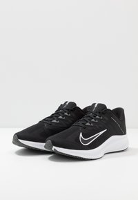 Zwarte sport schoenen met een bovenwerk van mesh, witte swoosh-logo, vetersluiting en een gestructureerde zwarte zool. Ontworpen voor comfort en ondersteuning.