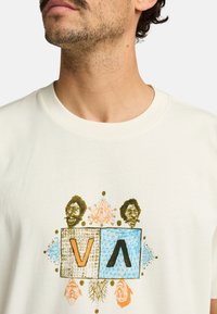 Kermanvärinen t-paita, jossa on graafinen printti, jossa esiintyvät "V", "A" ja abstrakteja kuvioita oranssina, sinisenä ja kultaisena. Pehmeä puuvillakangas.