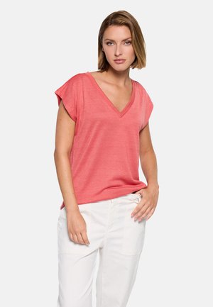 Mujer con cabello rubio corto, vistiendo una blusa coral de manga corta y escote en V, y pantalones blancos, de pie con una mano en el bolsillo frente a un fondo blanco.