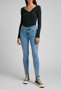 Top de manga larga negro con textura acanalada y botones en el frente, combinado con jeans ajustados azul claro y zapatillas altas verdes, presentado contra un fondo neutro.