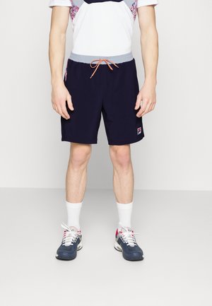 Homme portant un short à cordon de serrage bleu marine, des chaussettes blanches montantes, des baskets bleu marine et blanches, et un t-shirt blanc à motif, debout sur un fond uni.