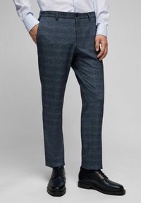 Dunkelblaue karierte Hose aus einem strukturierten Stoff, mit geradem Beinverlauf, Fronttaschen und Knopfverschluss. Getragen mit schwarzen Schuhen.